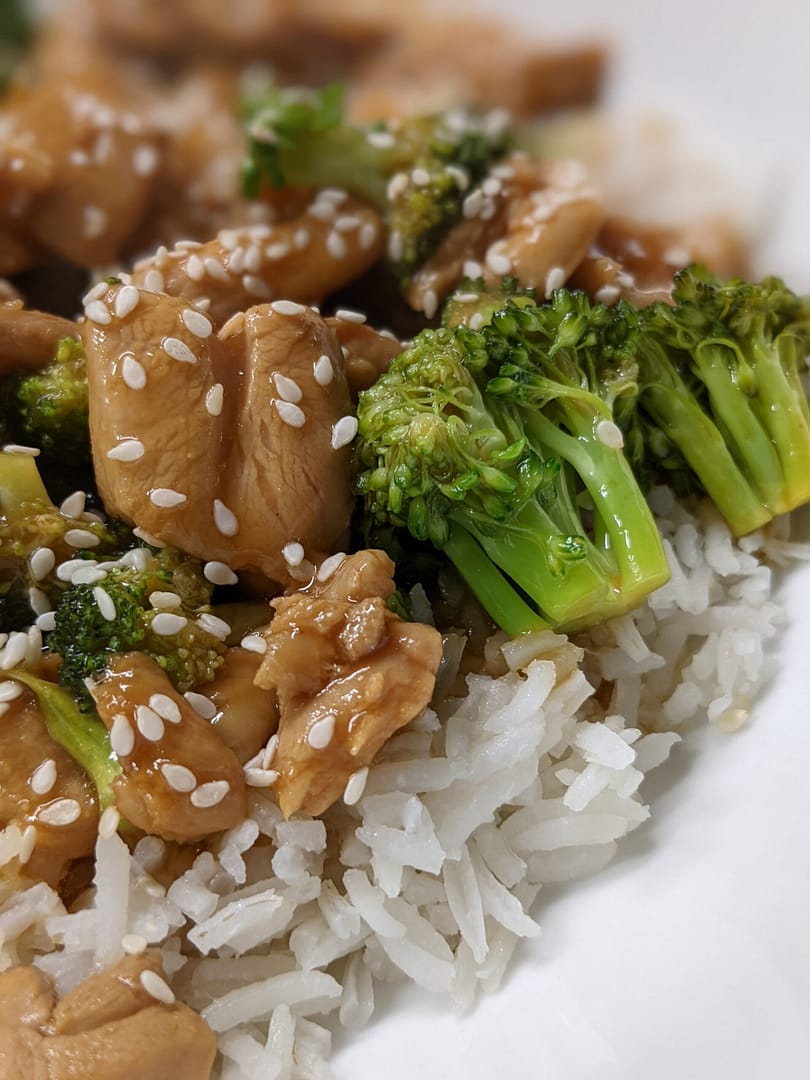 Close-up of homemade chicken and broccoli stir-fry served over fluffy white rice, topped with sesame seeds and coated in a savory soy-garlic sauce