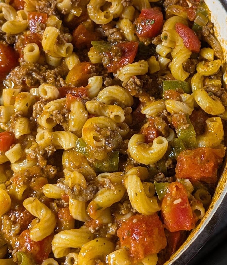 Close-up of homemade American Goulash in a red Dutch oven, featuring elbow macaroni, ground beef, green bell peppers, and diced tomatoes in a savory tomato-based sauce — a hearty one-pot comfort food.