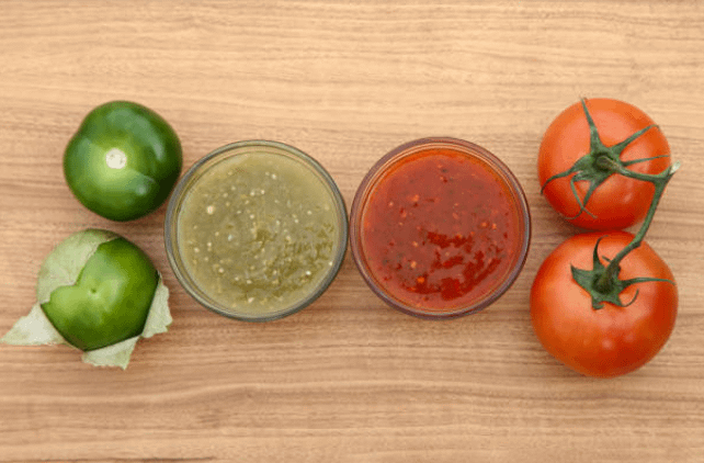 Red and green enchilada sauces in glass bowls, surrounded by fresh tomatillos and ripe tomatoes on a wooden surface, highlighting the key ingredients for traditional Mexican sauces.
