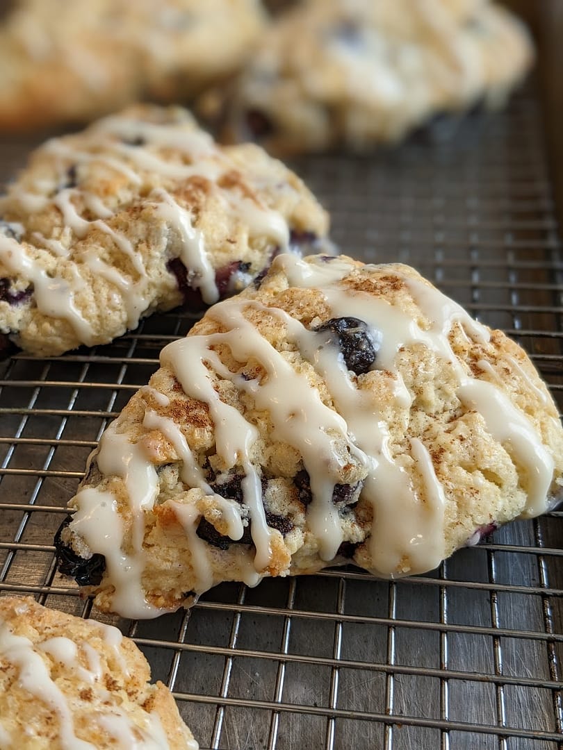 Close-up of a golden blueberry scone drizzled with creamy vanilla icing, resting on a cooling rack. The scone is buttery, flaky, and studded with juicy blueberries.