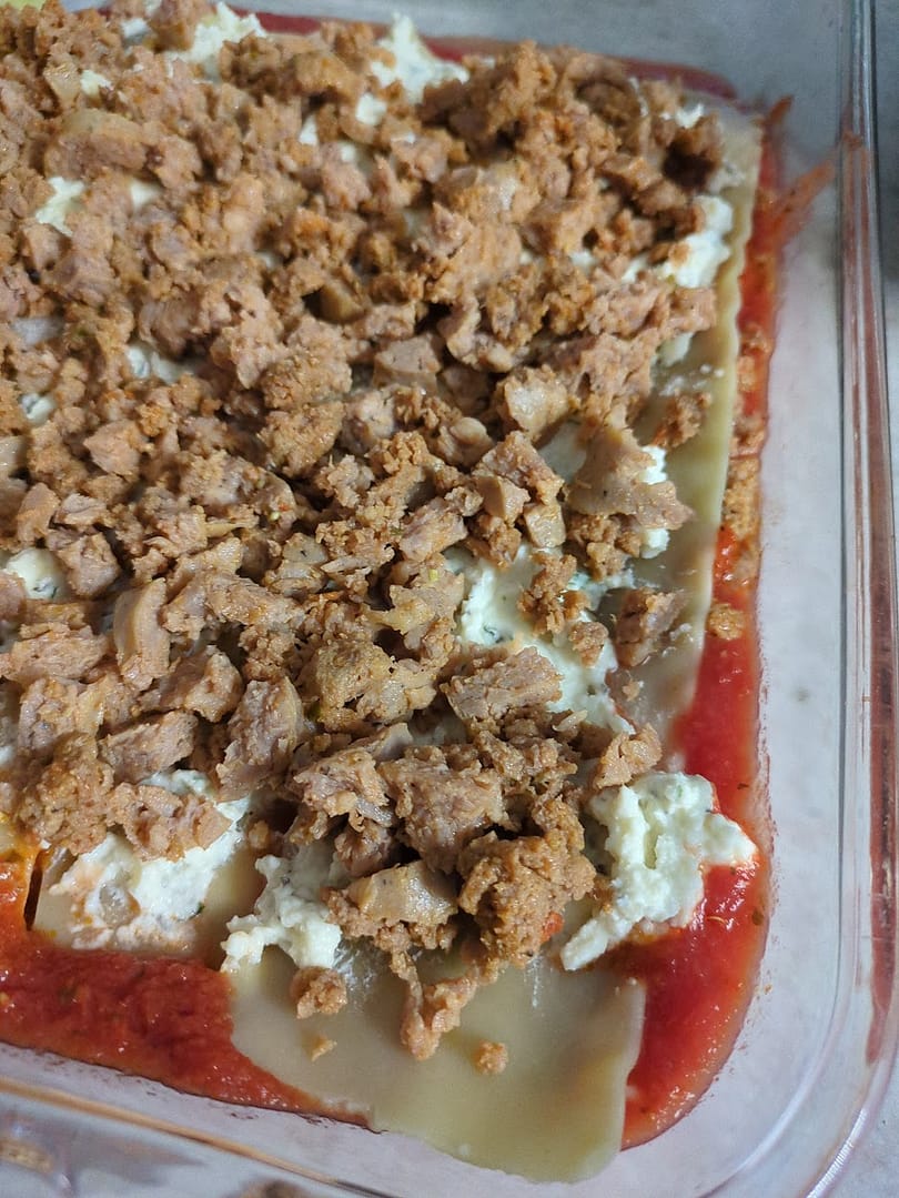 Layered lasagna in progress with lasagna noodles, ricotta mixture, and a generous topping of crumbled meatball and sausage filling in a glass baking dish.